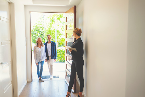 Real estate agent showing a young couple a new house. The house is contemporary. All are happy and smiling. The couple are casually dressed and the agent is in a suit. The couple are being greeted at the front door by the real estate agent. Copy space