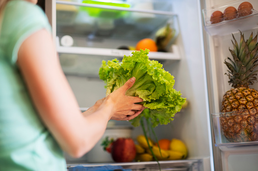 Unrecognizable female opens a refrigerator and takes green salad, female hands holding green salad in domestic kitchen.refrigerator with fruits and vegetables on background.