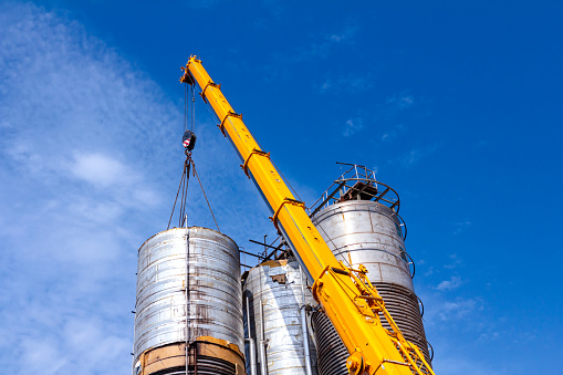 Crane is carry and keeps balance with heavy metal silo in industrial complex.