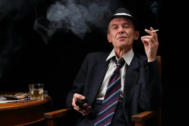Portrait of a gangster in retro style with a cigarette, a gun and a glass of whiskey on a dark background