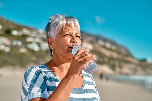 Cropped shot of an attractive senior woman drinking water from a bottle on the beach during the day