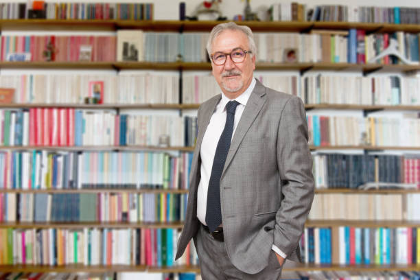 istock Senior teacher standing holding a book in front of a bookcase 1155879570