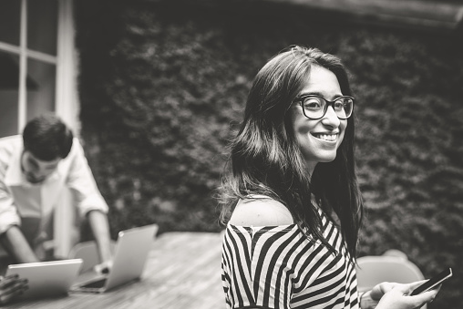 young woman with mobile standing in front of work desk outdoors , two working people in the background