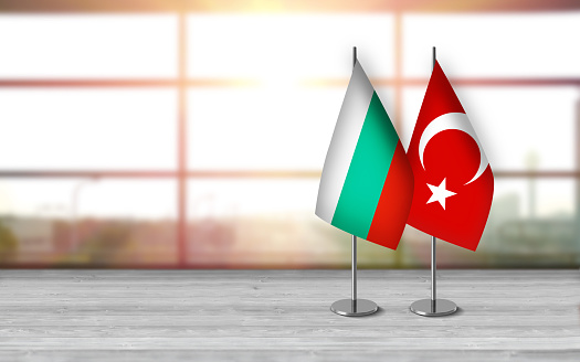 3D bulgarian and turkish flags with metallic pole, standing together on a white wooden desk in front of sunny window background. With large copy space you can write your own titles effectively. Also you can use this compositon as square in social media channels like instagram etc.