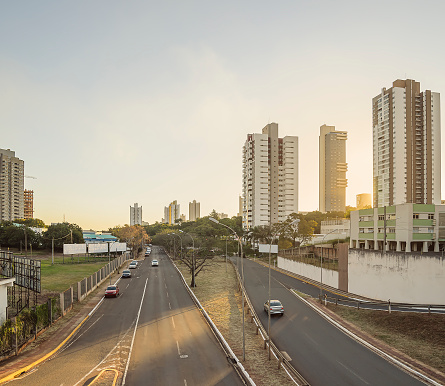 Two lanes avenue with few cars, trees, blank advertising billboards, and buildings around. Sunset at the large avenues of the capital city Campo Grande - MS, Brazil. Ricardo Brandao avenue.