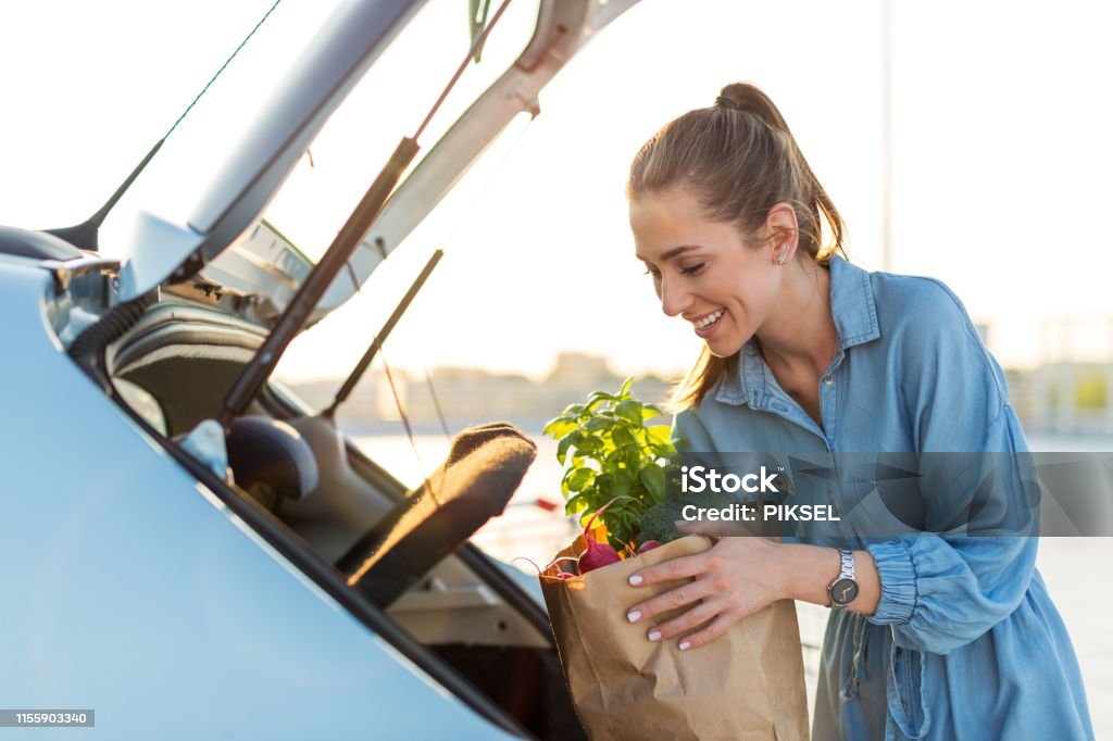 Happy girl packing groceries Young woman in car park carrying shopping bag of groceries Supermarket Stock Photo