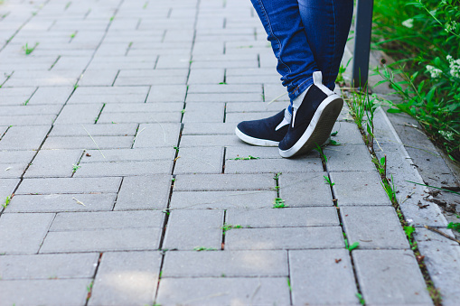 Young female with black modern sneakers and jeans standing with her feet crossed on a grey pavement with rectangle blocks – Person with trendy shoes on the sidewalk with copy space