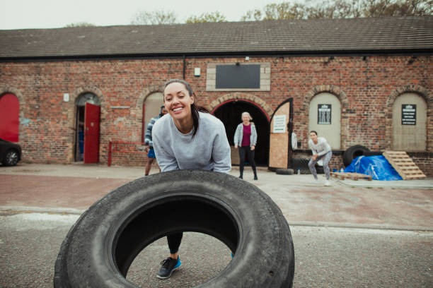 Woman flipping a tire outside of a gym gym. She is smiling and enjoying the activity. There are people behind her cheering her on.