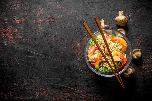 Instant noodles in a glass bowl with shrimp, vegetables and mushrooms. On dark rustic background