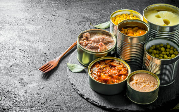 Various open tin cans of canned food on a stone Board with a fork. Various open tin cans of canned food on a stone Board with a fork. On black rustic background canned food stock pictures, royalty-free photos & images