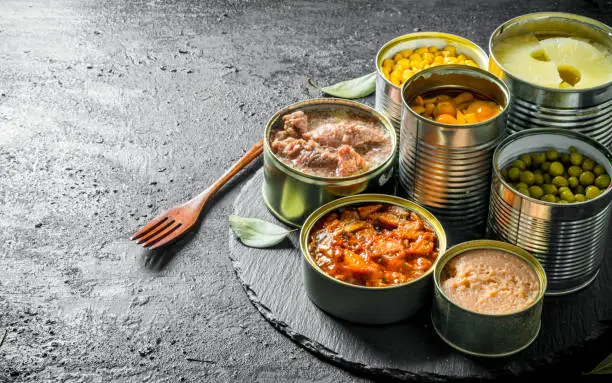 Various open tin cans of canned food on a stone Board with a fork. On black rustic background