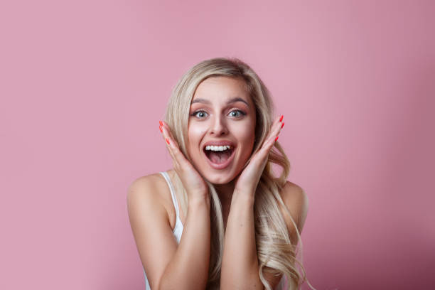 istock Surprised beautiful woman holds cheeks by hand on pink studio background 1155945311