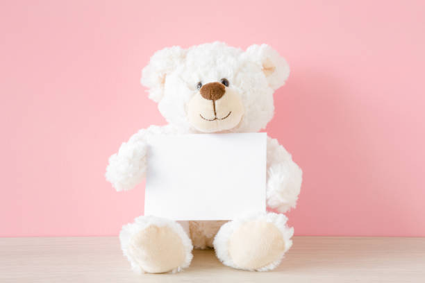 istock Smiling teddy bear sitting at pastel pink wall and holding white card. Empty place for inspiration, emotional, sentimental text, lovely quote or sayings for good mood. Front view. 1155952141