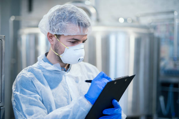 istock Technologist specialist with protective mask and hairnet holding clipboard checklist and writing production results. 1155960537