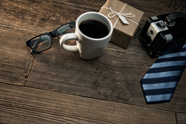 This is a close up photo of a cup of coffee, and old retro camera, a gift in brown paper and a blue striped tie on a wood table for Father's Day gifts and presents