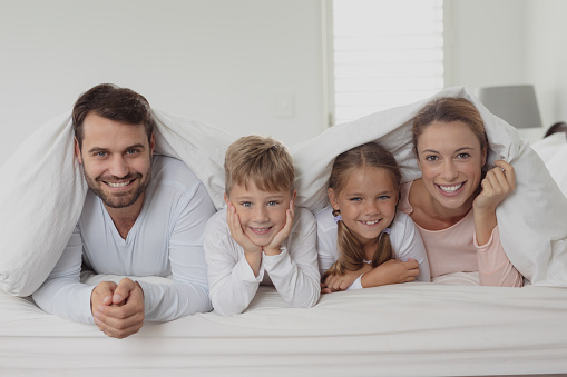 Front view of happy Caucasian family lying under blanket on bed in bedroom at home
