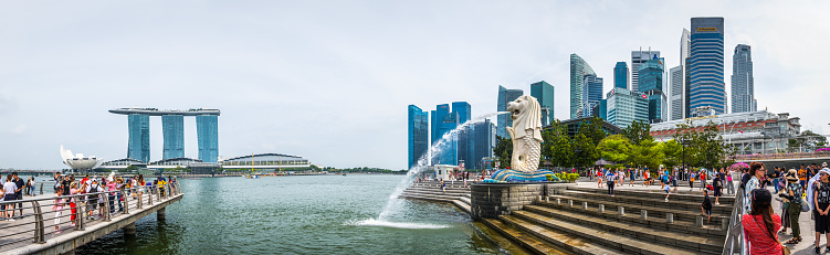Crowds of tourists on the waterfront beside the Merlion statue overlooked by the futuristic skyscraper cityscape of the Central Business District and Marina Bay Sands, Singapore.
