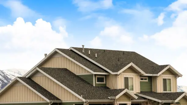 Panorama frame House exterior with view of the dark pitched roof against a cloudy blue sky. A mountain blanketed with snow can be seen behind the home.