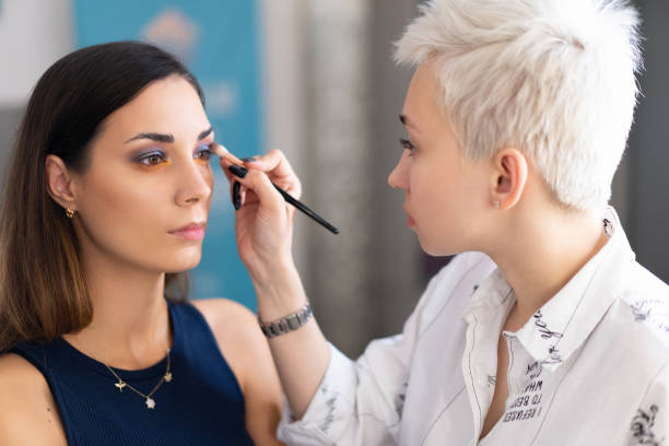 istock Make-up artist applying bright base color eyeshadow on model's eye and holding a shell with eyeshadow on background 1155985430