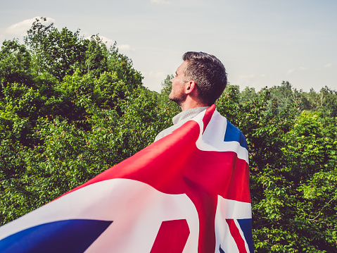 Attractive man waving a British Flag against a background of trees and blue sky. View from the back, close-up. National holiday concept