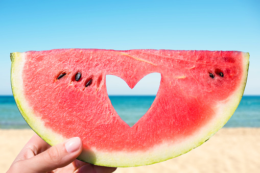 Ripe piece of watermelon with heart shape hole in female hands on the background of the beach on a hot summer day. Concept