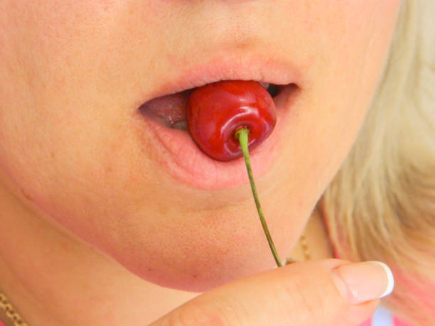 Woman bites and eats cherry closeup image. Part of face with mouth and lips. Red fruit is in fingers with clear nails. Isolated on white background Woman bites and eats cherry closeup image. Part of face with mouth and lips. Red fruit is in fingers with clear nails. Isolated on white background tasting cherry eating human face stock pictures, royalty-free photos & images