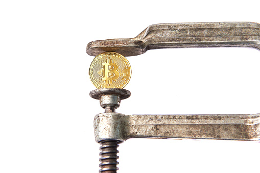 Salzburg, Austria - June 15, 2019: BITCOIN coin being squeezed in vice on white background; concept of cryptocurrency bitcoin under pressure. Prohibition of cryptocurrencies, regulations, restrictions or security. Isolated. Close up.