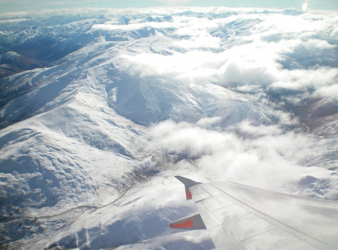 A view over the wing of a plane to snow-covered mountains. The plane is above some white clouds. The peaks of the mountains are just below the clouds.