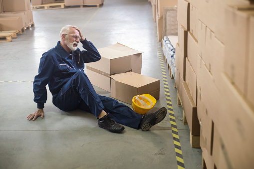 Warehouse worker sitting on a warehouse floor fallen from injury. About 60 years old, Caucasian senior male.