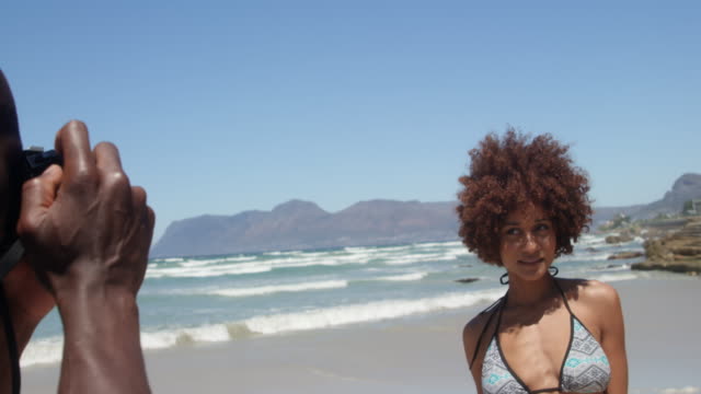 Side view of young African american man taking pictures of woman with digital camera on the beach 4k