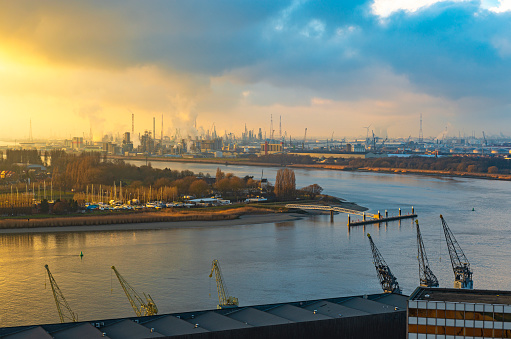 The second largest industrial harbor of Europe at sunset with cranes in the foreground, chemical and nuclear plants in the background and the Scheldt River, Antwerp, Belgium.
