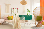 Peacock chair with olive green pillow in trendsetting bedroom interior with white metal bed with colorful pillows and handmade flower board and macrame