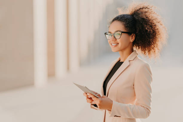 profil tiré de la femme à peau foncée heureux avec des cheveux afro, habillé avec élégance tient ordinateur tablette dans les promenades des mains à la rue porte des lunettes optiques connectées à internet haute vitesse. style de vie, technologie - standing digital tablet adult beautiful photos et images de collection