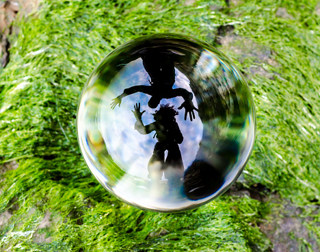 dark silhouettes of man and woman are distortedly reflected in glass ball lens lying on the green grass
