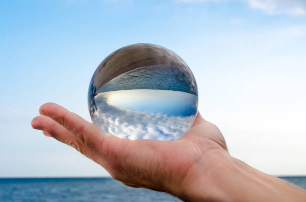 man's hand holds a glass orb lens in which an inverted reflection of the blue sea sky and clouds
