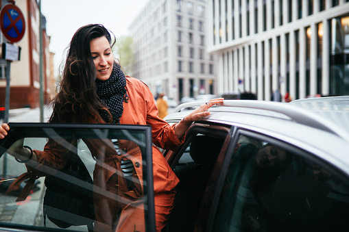 Young commuter woman catching a car ride share service in Berlin. She is smiling, wearing a fashionable coat, entering the vehicle.