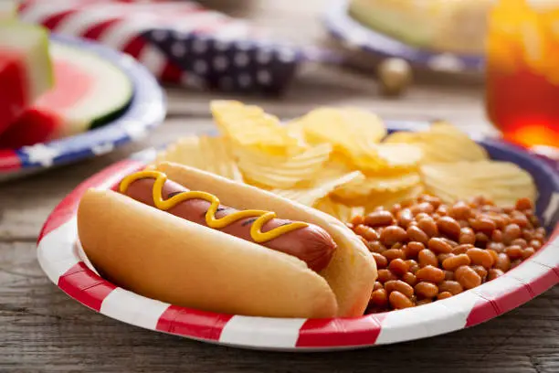 Forth of July or Memorial Day backyard barbecue with grilled hotdog, baked beans, potato chips, grilled corn watermelon, and iced tea on rustic wood picnic table.