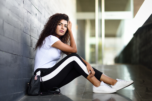 African woman with black curly hairstyle sitting on urban floor. Arab girl in sport clothes in the street.