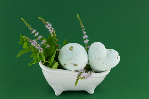 mint bath bombs.green bombs for the bath set with mint extract, sprigs of fresh blooming mint in a ceramic white bath on a dark green background.