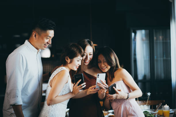 jeune homme asiatique restant à côté d'un groupe de jeune femme blottis ensemble et riant tout en regardant le smartphone pendant la partie - photographing smart phone friendship photo messaging photos et images de collection