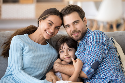 Portrait of happy young family with little kid relax sit on couch hugging looking at camera, smiling parents cuddle with preschooler daughter rest on cozy sofa at home posing for picture