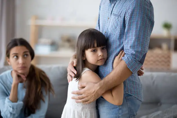 Sad little girl hug father upset by father leaving on business trip, unhappy young family in living room with preschooler daughter embrace say goodbye to father, parents divorce hurt small child