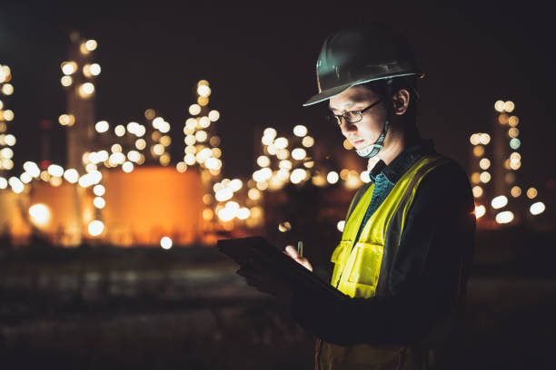 Asian man engineer using digital tablet working late night shift at petroleum oil refinery in industrial estate. Chemical engineering, fuel and power generation, petrochemical factory industry concept stock photo