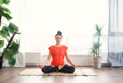 Attractive young brunette woman exercising and sitting in yoga lotus position while resting at home