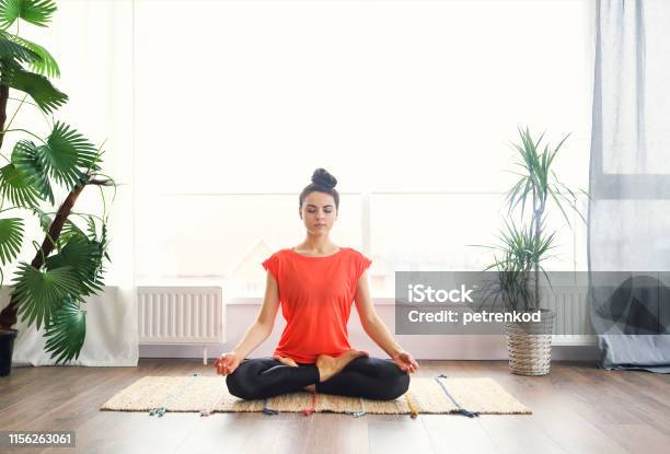 Attractive Young Woman Exercising And Sitting In Yoga Lotus Position While Resting At Home Stock Photo - Download Image Now