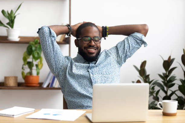 lächelnder afroamerikanischer geschäftsmann hält hände hinter kopf sitzend im büro - black break stock-fotos und bilder