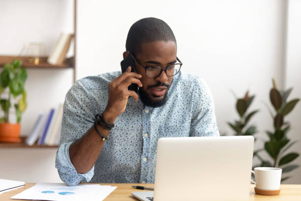 Serious african-american employee making business call focused on laptop screen Serious african-american employee making business call focused on laptop at workplace. Black businessman consulting customer, discussing financial report. Contract negotiation and discussion concept businessman african descent on the phone business person stock pictures, royalty-free photos & images