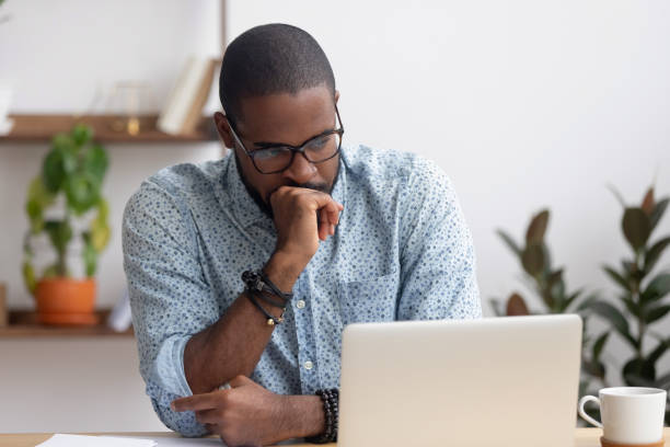 Head shot serious puzzled African American businessman looking at laptop Head shot serious puzzled African American businessman looking at laptop screen sitting in office. Executive managing thinking received bad news keeping fist at chin waiting hoping positive result skeptical look stock pictures, royalty-free photos & images