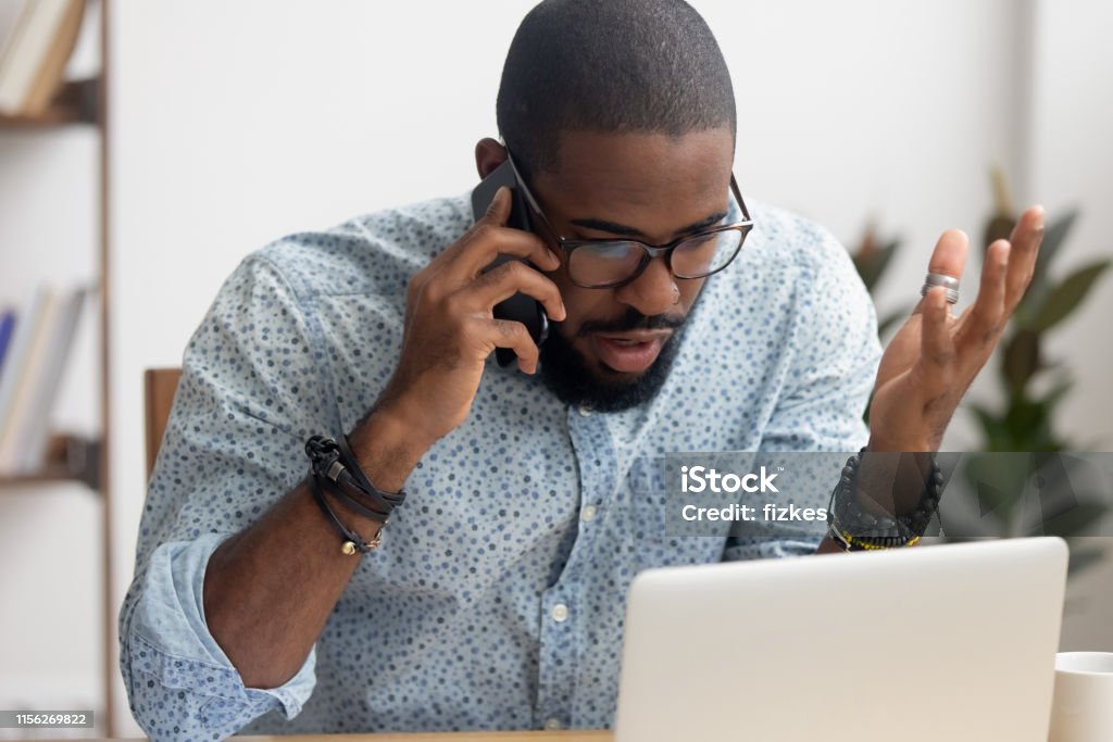 Angry mad african-american businessman talking on cellphone in office Angry mad african-american businessman talking on cellphone looking at laptop in office. Shocked manager received bad news discussing failing profit statistics with coworker, solving problem distantly Customer Stock Photo