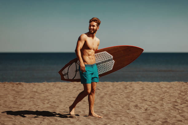 A mature man, in his 40s, at the beach carrying a fish surfboard.  He is  standing on the sand, ready to get in the water and go surfing, shading his eyes from the sun with his hand, looking out over the water.